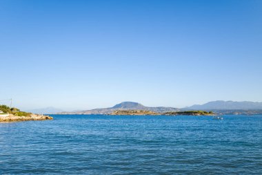 Sakin mavi sular, Girit adasında Marathi yakınlarında berrak bir gökyüzü altında küçük adaları ve puslu dağları olan engebeli bir kıyı şeridine doğru uzanır..