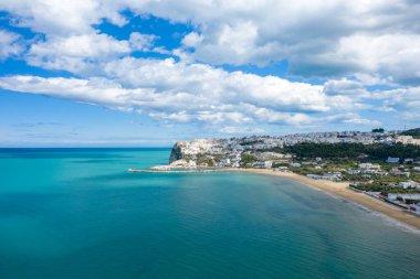 Peschicis 'in kumlu körfezi ve turkuaz denizi süpürdüğü geniş bir hava manzarası. Burnun altında dağınık bulutlarla dolu beyaz binalar kümelenmiş..