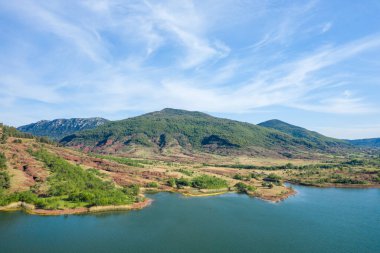 Parlak bir gökyüzünün altında yuvarlanan yeşil tepeler, kırmızı toprak ve berrak mavi suyun yer aldığı Salagou Gölü 'nün hava manzarası. Manzara doğal dokuları ve sakin, ferah bir atmosferi gösteriyor..