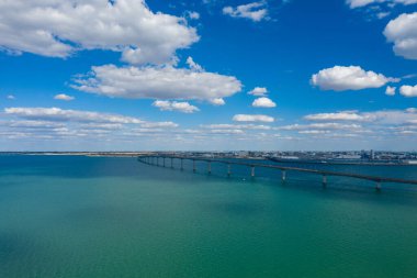 Uzun beton köprü, gökyüzü altında kümülüs bulutlarıyla dolu, Charente-Maritime 'deki Ile de Re' yi anakaraya bağlayan canlı turkuaz suları kapsar. Geniş görünüm batı bölgesinin açık kıyı manzarasını ve sakin atmosferini vurguluyor.