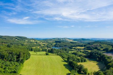 Yeşil tarlalar, sık ormanlık alanlar ve Morvan bölgesindeki kayan tepelerin arasına yerleşmiş sakin bir göl. Manzara parlak mavi bir gökyüzünün altında ince bulutlarla aydınlık ve geniştir..