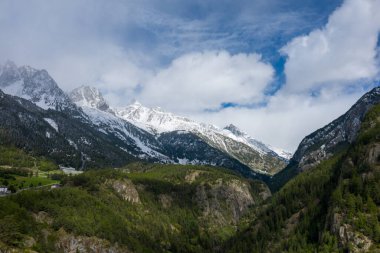 İtalya 'nın Val dAoste kentindeki yoğun yeşil ormanlar ve kayalık yamaçların üzerinde karla kaplı zirveler yükselir. Sahnede desenli yamaçlar, dağınık bulutlar ve Alpler 'de canlı bir bahar atmosferi yer alıyor..
