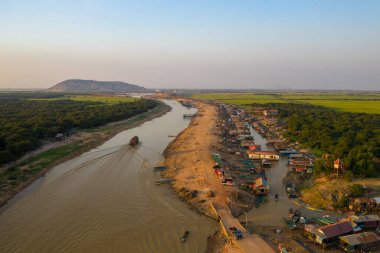 Kamboçya 'daki Tonle Sap' ın yakınlarındaki ıssız evleri ve tekneleri olan kırsal bir köyün havadan görünüşü. Manzarada dolambaçlı çamurlu bir nehir, yoğun yeşil orman ve açık akşam ışığı altında uzak bir tepe yer alır..