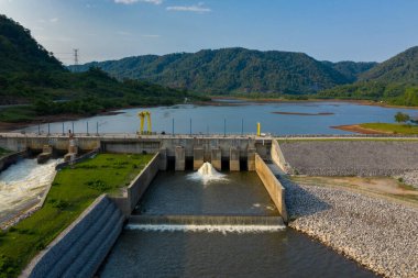 Geniş bir nehir boyunca uzanan modern bir hidroelektrik baraj, ormanlık tepeler ve kayalık setlerle desteklenmiş bir rezervuarla ayakta duruyor. Sahnede geometrik çizgiler, akan su ve mühendislik yapısı ile doğal manzara arasındaki zıt dokular yer alıyor.