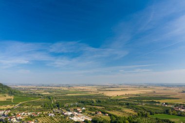 Geniş arazi manzarası ve yeşil çitlerle, bahçelerle çevrili kırsal bir köy ve engin, berrak bir mavi gökyüzünün altında açık tarım arazisi. Manzarada yumuşak tepeler, ekili topraklar ve Lorraine bölgesinde dağınık evler yer alır..