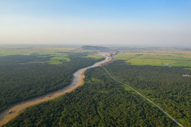 Kamboçya 'daki Tonle Sap yakınlarında yoğun yeşil orman ve pirinç tarlaları boyunca kıvrılan çamurlu bir nehrin havadan görünüşü. Geniş arazide uzak tepeler, açık kırsal alanlar ve berrak mavi bir gökyüzü var. Bu da bir huzur ve doğal hava hissi uyandırıyor.