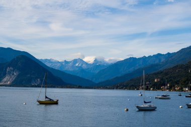 Kuzey İtalya 'daki Maggiore Gölü' nün sakin sularında dinlenen birçok yelkenli engebeli dağlarla çevrili ve yumuşak bulutlarla kaplı bir gökyüzü. Sakin göl manzarasında uzak tepeler, ormanlık tepeler ve huzurlu bir rıhtım manzarası yer alıyor.