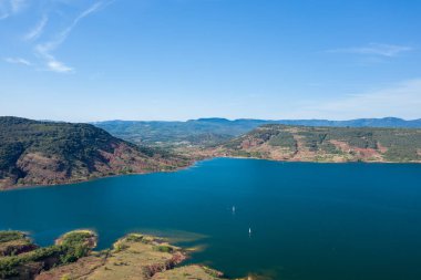 Lac de Salagou 'nun yemyeşil tepelerle çevrili derin mavi suları ve berrak gökyüzünün altındaki engebeli kırmızı yamaçlarıyla geniş bir hava manzarası. Sahne sakin ve açık, uzak dağlar ve doğal alan hissi var..