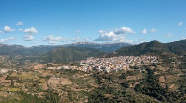 Sardunya 'daki parlak mavi gökyüzünün altında yuvarlanan tepeler, yamalı tarlalar ve dramatik dağ sırtları arasında yuva yapmış Orgosolo köyünün geniş açılı manzarası..