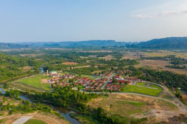Kırmızı çatılı evleri olan planlanmış bir yerleşim yeri, yeşil tarlalar ve açık kırsal alanlarla çevrili dolambaçlı bir nehrin yanında yer alıyor. Manzarada dağınık ormanlık, uzak dağlar ve açık gün ışığı altında bir tarım arazisi parçası yer alıyor..