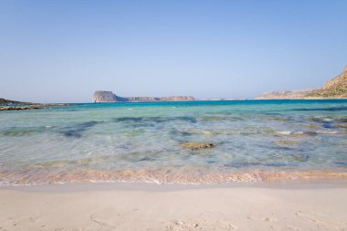 Girit 'in Balos kenti yakınlarındaki sakin bir sahilde, açık ve sığ dalgalar, açık mavi gökyüzünün altındaki uzak uçurumların üzerinde süzülüyor. Sahne açık, güneşli ve huzurlu..