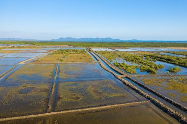 Hava perspektifi, Kamboçya 'nın Marais tuzlu sularındaki su basmış tuz buharlaştırma havuzları ve yeşil bitki örtüsünü ortaya çıkardı. Sahnede geometrik desenler, toprak dokular ve berrak gökyüzünün altındaki uzak mavi dağlar yer alıyor..