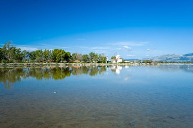 Sakin su, Yunanistan 'ın batısındaki Tourlida lagünündeki ağaçların ve beyaz bir kilisenin ufukta görünen uzak dağlarla dolu canlı mavi bir gökyüzünün altındaki yansımasını yansıtıyor. Sakin sahne berrak yansımalar, yemyeşil ve açık sulak alan içerir..