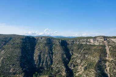 Navacelles Sirki 'ndeki engebeli kireçtaşı uçurumlarının ve derin kanyonun havadan geniş perspektifi. Güneş ışığı desenli yamaçlara vurgu yapıyor ve canlı bir gökyüzüne karşı seyrek yeşillik sergiliyor..