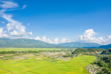 Kırsal bir köy ve vadi boyunca uzanan yemyeşil pirinç tarlalarının geniş hava manzarası ve canlı mavi gökyüzü altında yuvarlanan dağlar ve Dien Bien Phu yakınlarında dağınık beyaz bulutlar. Parlak güneş ışığı canlı renkleri ve açık manzarayı güçlendirir..