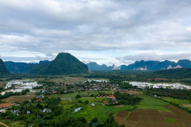 Yeşil pirinç tarlaları ve Vang Vieng, Laos yakınlarındaki dramatik kireçtaşı tepeleriyle çevrili bir köyün havadan görüntüsü. Bulutlu gökyüzü yemyeşil kırsal bölgelerin üzerine yayılarak karst oluşumlarını ve tarlaları vurguluyor..