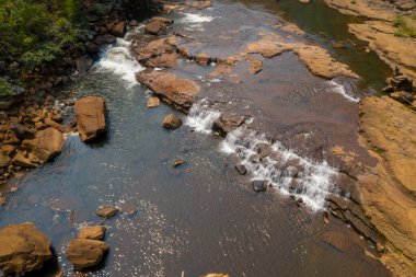 Hava perspektifi, düz taş çıkıntıların ve güney Laos 'taki Tad Tayicsua yakınlarındaki nehirdeki dağılmış kayaların üzerinden akan yumuşak bir çağlayanı gösteriyor. Sıcak gün ışığı, bu sakin tropikal ortamda toprak dokuları ve ince dalgalanmaları vurgular..