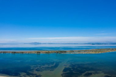 Yunanistan 'ın batısındaki Missolonghi tuz gölünün geniş manzarası, sakin mavi su, dar bir arazi şeridi ve berrak bir gökyüzünün altındaki uzak sıradağlar. İnce yansımalar ve geometrik çizgiler sakin ve açık bir manzara yaratır..