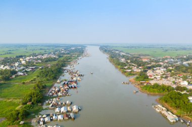 Vietnam 'daki Chau Doc nehrinde yüzen bir köye geniş bir hava perspektifi, su yolu boyunca yığınla stilt ev ve tekne bulunuyor. Görüntü, yemyeşil nehir kıyılarını, açık kırsal alanları ve berrak mavi gökyüzünün altındaki geniş pirinç tarlalarını yakalıyor..