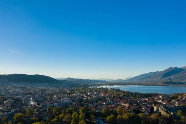 Panoramik bir hava perspektifi, kırmızı kiremitli çatılar, bereketli ağaçlar ve Ioannina Gölü 'nün geniş mavi suları ile çevrili İoannina şehrini ve Pindus Dağları' nı ortaya çıkarır. Olay yeri gün ışığında yıkanıyor.