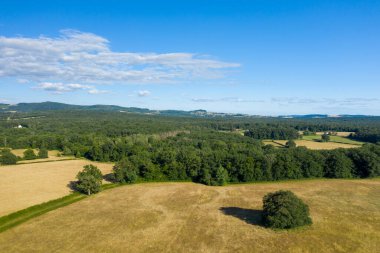 Morvan bölgesinde yoğun yeşil ormanlarla çevrili altın yaz otlaklarının geniş hava manzarası, kayan tepeler ve bulutlarla kaplı parlak mavi bir gökyüzü. Manzara, açık kırsal dokulara ve huzurlu, güneşli bir havaya sahiptir..
