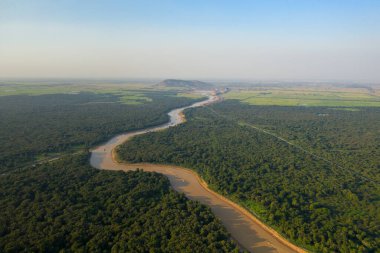 Kamboçya 'daki Tonle Sap yakınlarındaki yoğun yeşil ormanı ve açık tarım arazisini kesen dolambaçlı bir nehrin havadan görünüşü. Geniş arazide zengin bitki örtüsü, uzak tepeler ve kırsal kesimin sakin atmosferini yakalayan açık mavi bir gökyüzü bulunur.