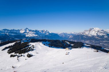 Megeve 'deki çam ağaçları ve dağ kireçleriyle benekli karlı tepe manzarası, dramatik alp zirveleri ve açık mavi bir kış atmosferi oluşturan derin mavi gökyüzü..