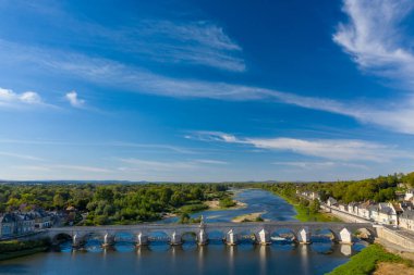 La Charite sur Loire 'deki Loire Nehri' ni kaplayan tarihi bir taş köprünün geniş bir manzarası. Geleneksel evler nehir kıyısını ve yemyeşil alanları kaplıyor. İnce bulutlar parlak mavi bir gökyüzünde sürükleniyor.