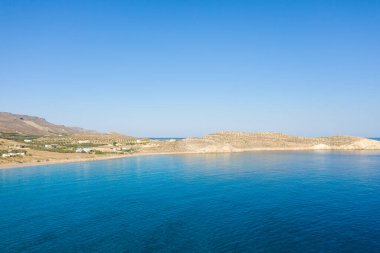 Girit, Ambelou 'nun kumlu körfezi ve engebeli tepeleriyle canlı, bulutsuz bir gökyüzünün altında buluşan berrak mavi suyun geniş bir görüntüsü. Sakin sahne ince dokular ve nazik doğal renkler içerir..