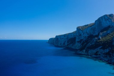Baunei, Sardunya 'daki Cala Goloritze yakınlarında Akdeniz' in derin safir sularının üzerinde yükselen kireçtaşı kayalıkları. Açık gökyüzü ve parlak mavi renkler sakin ve geniş bir kıyı manzarası yaratır..