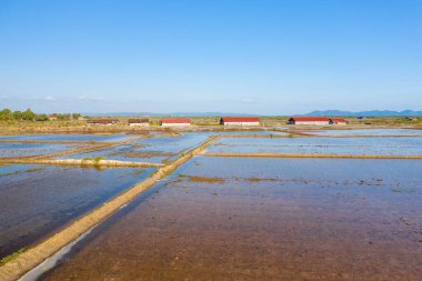 Kamboçya, Kamboçya yakınlarındaki geniş su basmış tuz buharlaştırma göletlerinin kenarında kırmızı çatıları olan kırsal tuz depolama barakaları. Manzara geometrik su desenleri, açık gökyüzü ve uzak mavi dağlar içerir, sakin atmosferi yakalar.