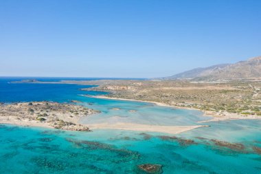 Geniş kumlu lagünü, sığ turkuaz suları ve bulutsuz mavi gökyüzünün altındaki engebeli tepeleriyle Girit 'teki Elafonisi plajının geniş hava manzarası. Sahne parlak ve davetkar, Akdeniz kıyı güzelliğini gözler önüne seriyor.