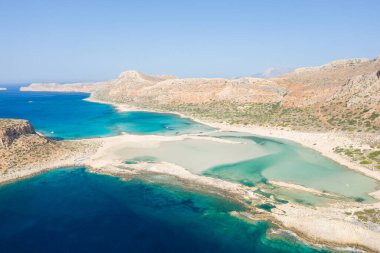 Yunanistan 'ın Girit kentindeki Balos Sahili' nin sığ turkuvaz suları, kumlu körfezleri ve parlak bulutsuz bir gökyüzünün altındaki dramatik kayalık tepeleriyle hava manzarası. Sahne açık ve güneşli. Dokunulmamış doğal bir güzelliği çağrıştırıyor..