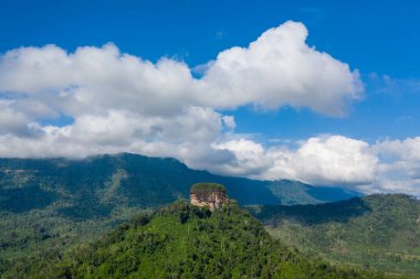 Yoğun tropikal ormanın üzerinde tek bir kayalık yükselir, yuvarlanan tepelerle çevrilidir ve büyük, dramatik bulutlarla dolu bir gökyüzünün altında canlı yeşilliklerle kaplıdır. Sahne, kırsal kesimin doğal dokularını ve katmanlı bitkilerini yakalıyor.