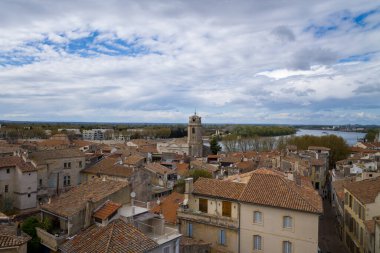 Arles 'in panoramik şehir manzarası, desenli terracotta çatıları, tarihi bir taş kulesi, ve Rhone Nehri dramatik, bulutlarla dolu bir gökyüzü altında sallanıyor..