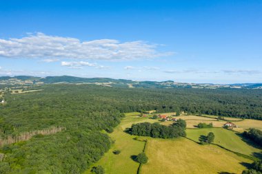 Morvan bölgesindeki küçük çiftlik evleri ile benekli, yoğun yeşil ormanlık ve açık arazilerin yer aldığı hava manzarası, dağınık bulutlu, parlak mavi bir gökyüzünün altında. Sahne, kırsal sükuneti, doğal dokuları ve yumuşak yaz ışığını taşır..
