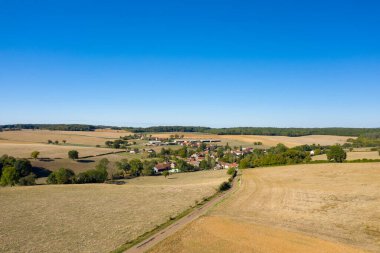 Hava perspektifi, Fransa 'nın Nievre bölgesinde geniş altın tarlalar, dağınık ağaçlar ve açık mavi gökyüzünün altındaki uzak ormanlık alanlarla çevrili küçük bir kırsal köy yakalar. Açık kırsal alan ve nazik ufuk bir duyguyu çağrıştırıyor