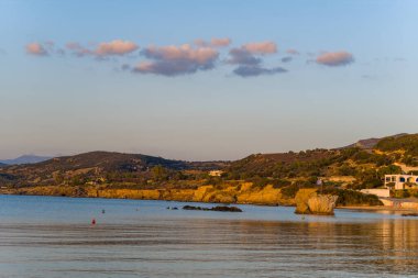 Sıcak güneş ışığı, Archangelos yakınlarındaki kıyı şeridi boyunca kayalık kayalıkları ve sakin suyu aydınlatır. Birkaç dağınık bulut ve uzak tepeler, huzurlu bir Akdeniz manzarası yaratır..