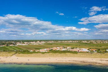 Aerial view of a whitewashed village with orange roofs bordered by farmland and sandy beach along the coast of Ile de Re, Charente-Maritime. The scene features clear blue sky, scattered clouds, and a tranquil shoreline atmosphere.