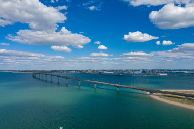 Ile de Re köprüsünü anakaraya doğru uzanan mavi-yeşil Atlantik sularına doğru uzanan geniş bir hava manzarası, dağınık kümülüs bulutlarıyla dolu bir gökyüzü. Kompozisyon açık kıyı manzarasını ve sakin atmosferi vurgular.