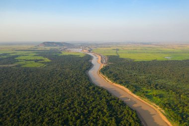 Yoğun orman ve Kamboçya 'daki Tonle Sap yakınlarında uzanan çamurlu bir nehrin havadan bakış açısı. Geniş arazide uzak tepeler, kırsal yerleşimler ve berrak mavi gökyüzü bulunur.