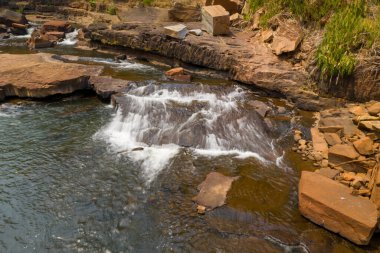 Temiz su, Laos 'un güneyindeki Tad Tayicsua yakınlarında açısal kayalar ve topraksı dokularla çevrili yumuşak bir şelale oluşturarak alçak, düz bir kaya çıkıntısının üzerinden akar. Sıcak gün ışığı, bu sakin nehirde kırmızımsı kahverengi taşları ve ince dalgaları vurgular.