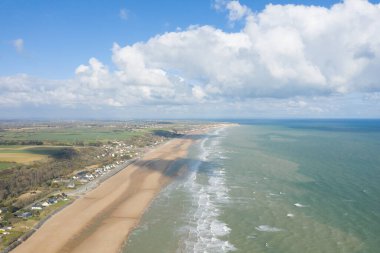 Yüksek drone perspektifi, Omaha Sahili 'nin yumuşak dalgalar, yeşil alanlar ve dramatik kümülüs bulutlarıyla dolu gökyüzünün altında dağınık evler ile çevrili uzun kumlu bölümünü yakalar..