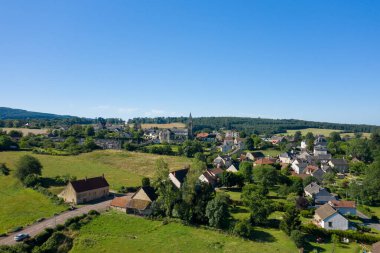 Saint-Honore-les-Bains 'in panoramik hava manzarası, bir grup geleneksel ev, belirgin bir kilise kulesi, ve canlı mavi gökyüzü altında yemyeşil alanlar.