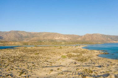 Geniş kumlu kıyı şeridi, seyrek bitki örtüsü, Girit 'teki Elafonisi' de bulutsuz bir gökyüzünün altında engebeli dağ sırtlarına kurulmuş berrak mavi sulara doğru uzanıyor. Yumuşak gün ışığı doğal dokuları ve sakin atmosferi güçlendirir..