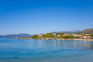 Huzurlu bir sahil köyü, sakin turkuaz su ve parlak mavi bir gökyüzünün altındaki uzak sıradağlarla çerçevelenmiş kayalık bir kıyı şeridi boyunca yer alır. Güneş ışığı yeşil ve geleneksel mimariyi vurguluyor ve sakin bir Akdeniz atmosferi yaratıyor.