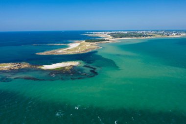 Turkuaz ve Quiberon yakınlarındaki derin mavi sulara uzanan kumlu bir tükürüğün geniş hava manzarası, görünür kaya çıkıntıları ve berrak bir gökyüzünün altında uzak bir kıyı şeridi. Sahne güneşli, açıklık ve kıyı sükunetini çağrıştırıyor..