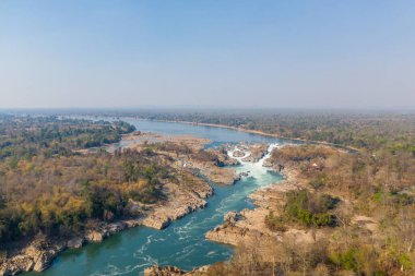 Güney Laos 'taki Mekong Nehri üzerindeki Khone Şelalesi' nin hava perspektifi, engebeli kayalık kanallardan akan beyaz su akıntıları. Kuru orman ve geniş nehir ovaları bulutsuz bir arazinin altında çarpıcı bir doğal manzara yaratır.