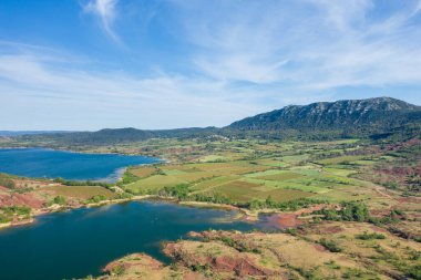 Lac de Salagou 'nun canlı mavi suları, yamalı tarımsal alanları ve parlak gökyüzünün altındaki engebeli tepeleri. Manzara zıt dokulara ve canlı doğal renklere sahiptir..