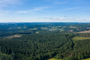 Yoğun çam ağaçlarından oluşan hava manzarası Morvan bölgesindeki yuvarlak tepelerin arasına yerleştirilmiş küçük bir köye doğru uzanıyor. Manzara, geniş mavi bir gökyüzünün altında, dağınık bulutlarla güneş ışığı ve canlı..